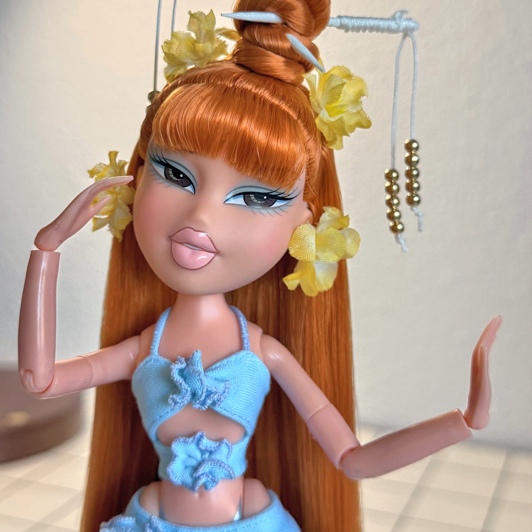 Close-up custom reroot example of Safari Auburn saran doll hair on Bratz Pop Starz Jade by @thedolldaddie. Medium shot highlighting the straight hair length, blunt bangs, high topknot bun with hair chopsticks, and yellow flower accessories.