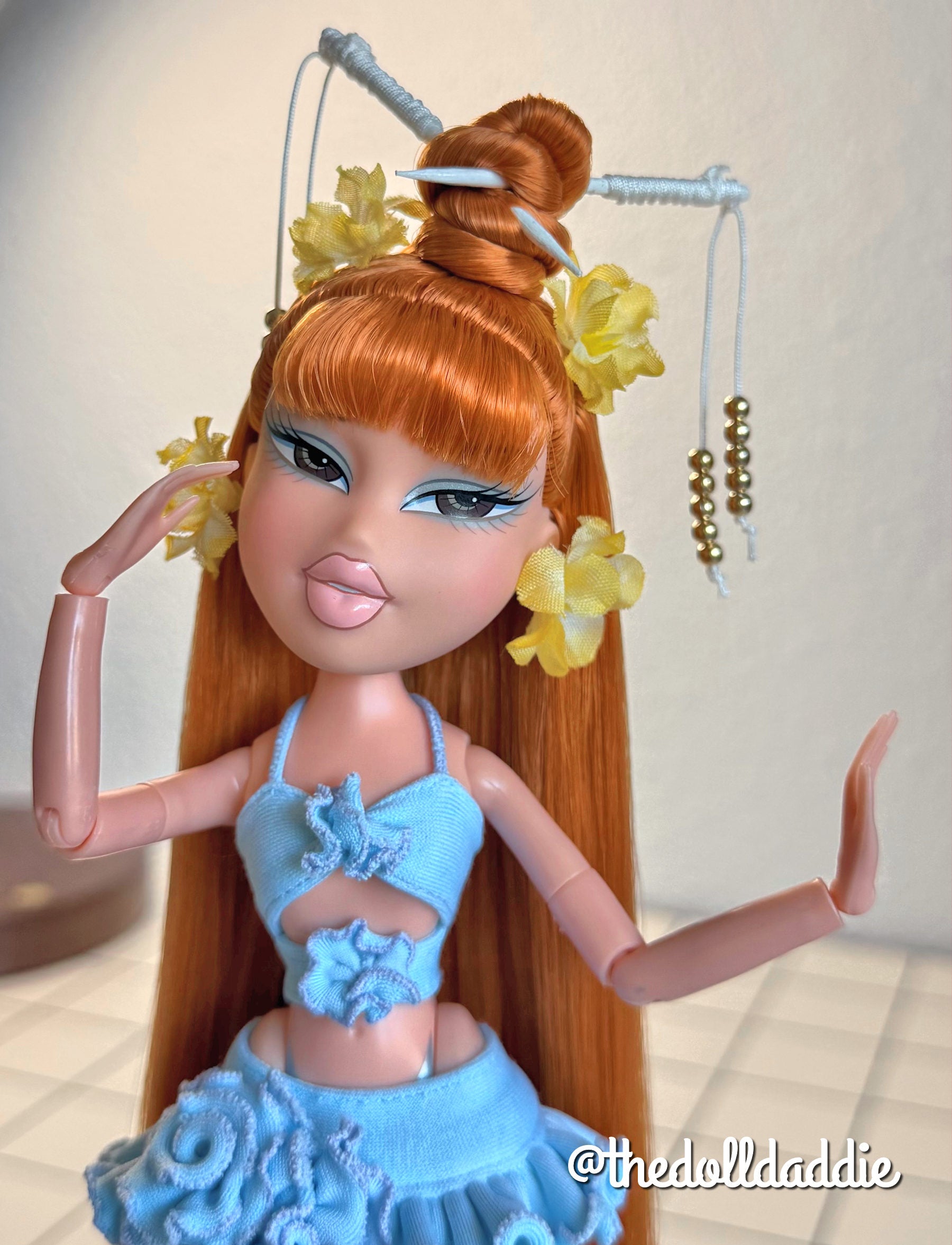 Close-up custom reroot example of Safari Auburn saran doll hair on Bratz Pop Starz Jade by @thedolldaddie. Medium shot highlighting the straight hair length, blunt bangs, high topknot bun with hair chopsticks, and yellow flower accessories.